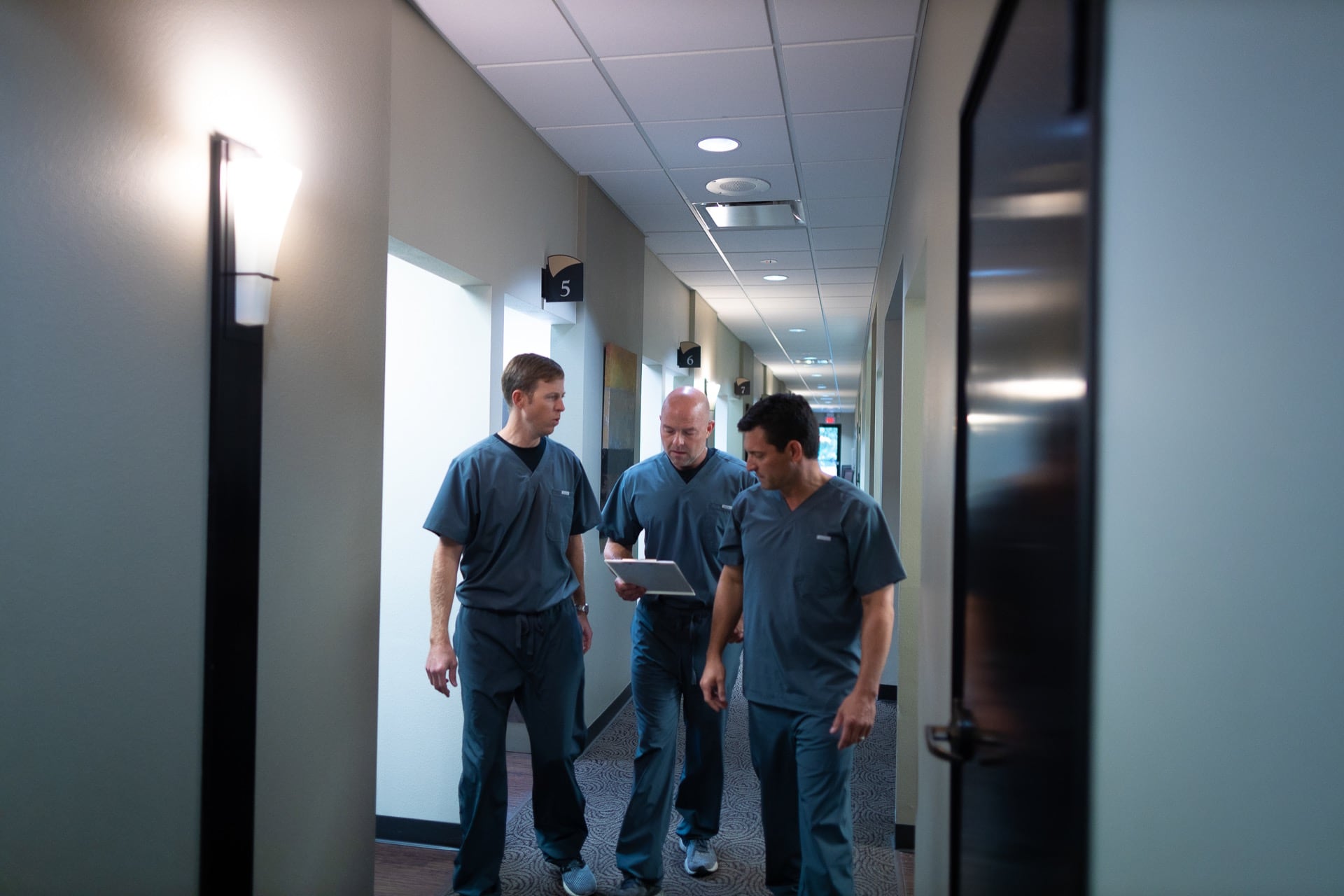 Three men in blue uniforms walking through a modern office hallway with fluorescent lighting.