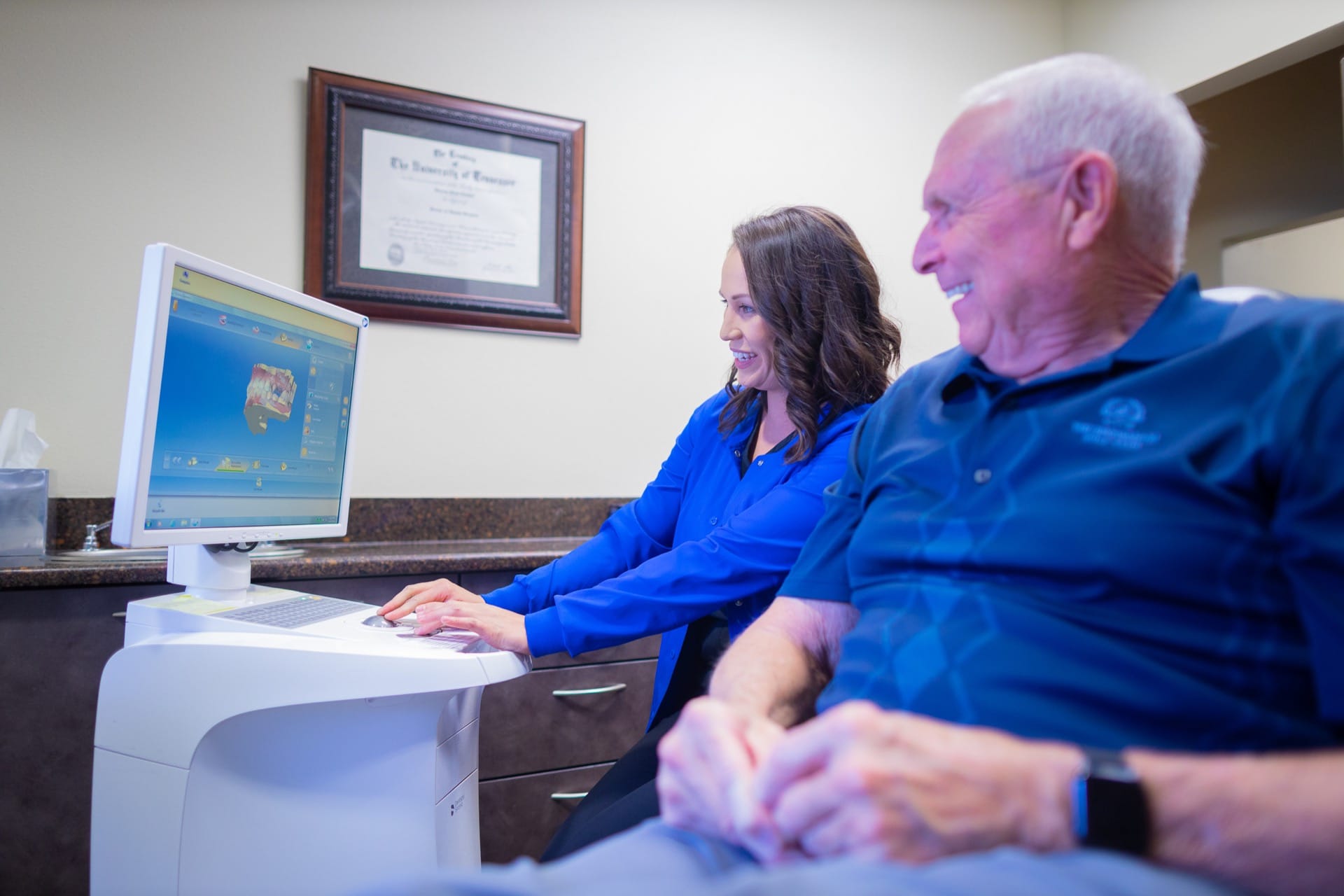 Two people in blue shirts working at computer in office with framed certificates on wall.