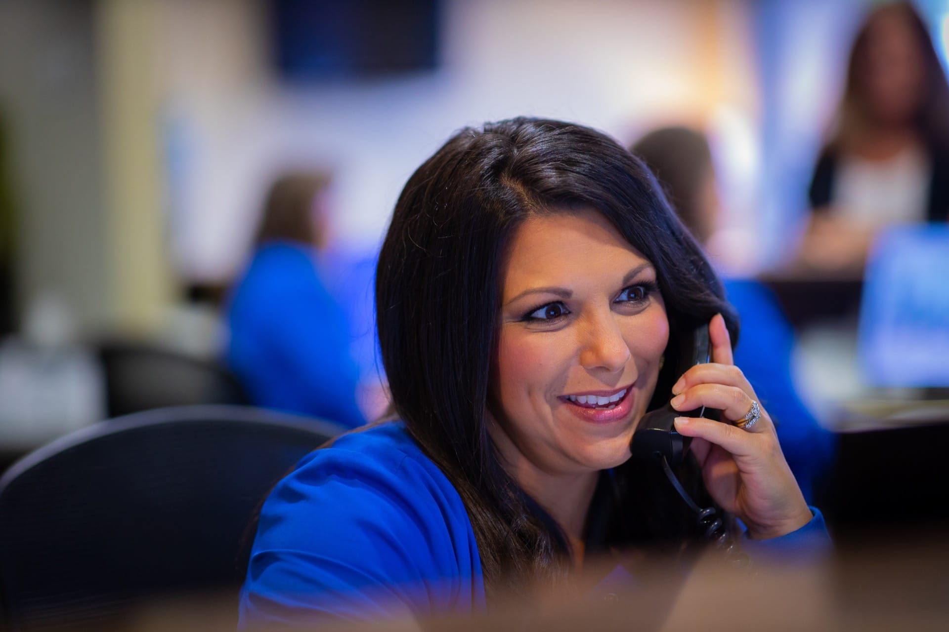 Woman in blue shirt smiling while talking on phone at office desk with colleagues in background.