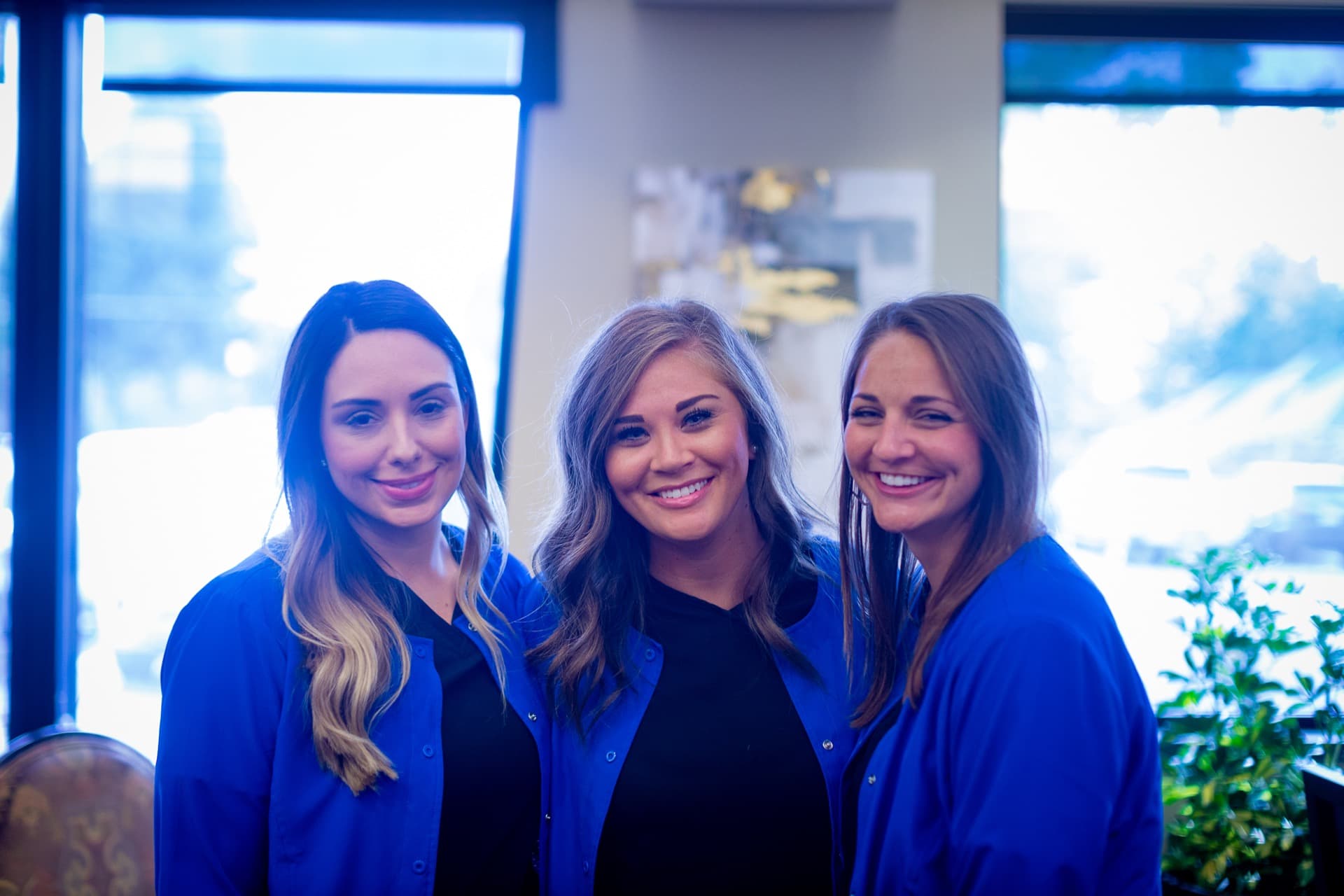 Three women in blue outfits smiling at camera in modern office setting with windows.