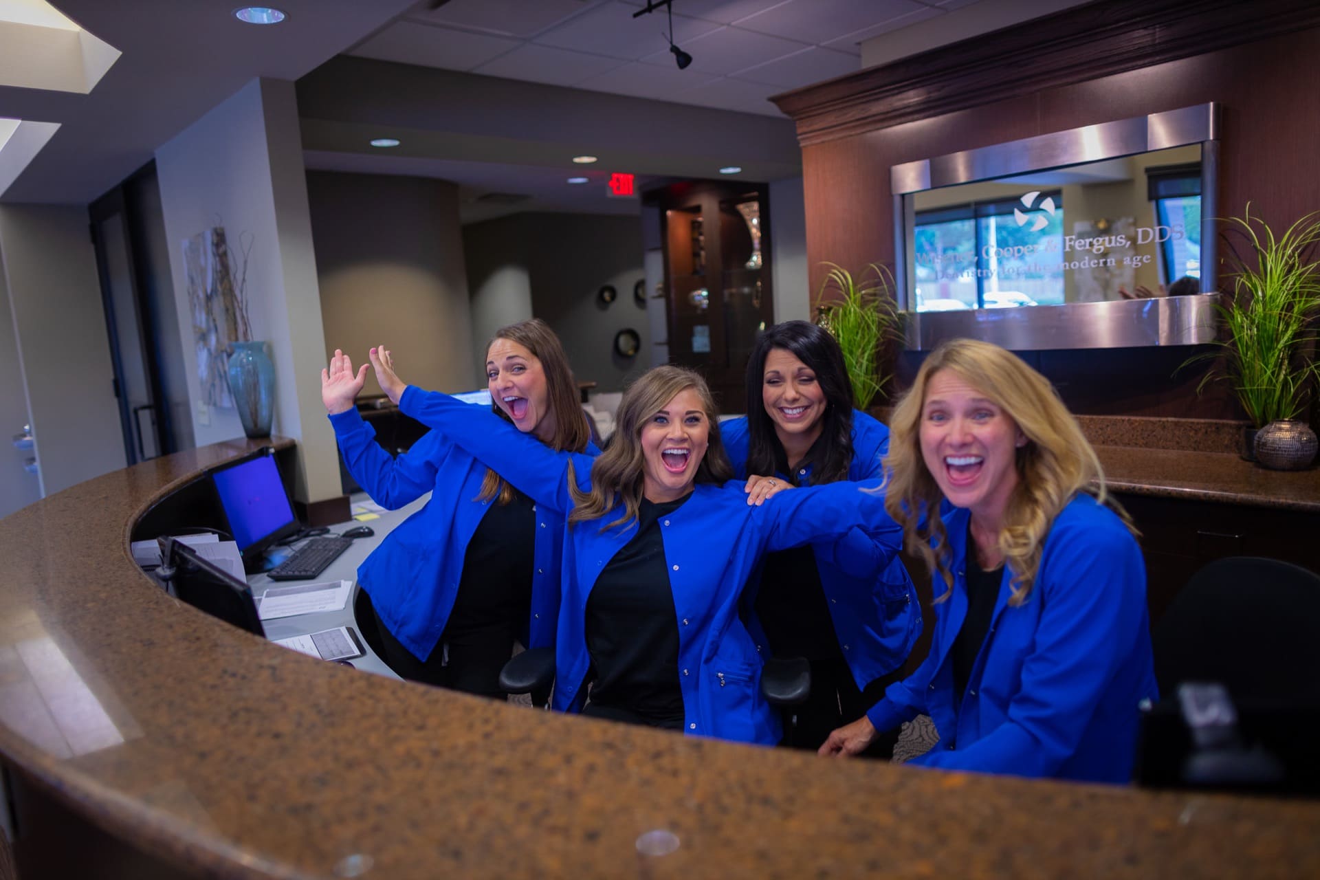 Team of five women in blue uniforms standing behind reception desk in modern office lobby.