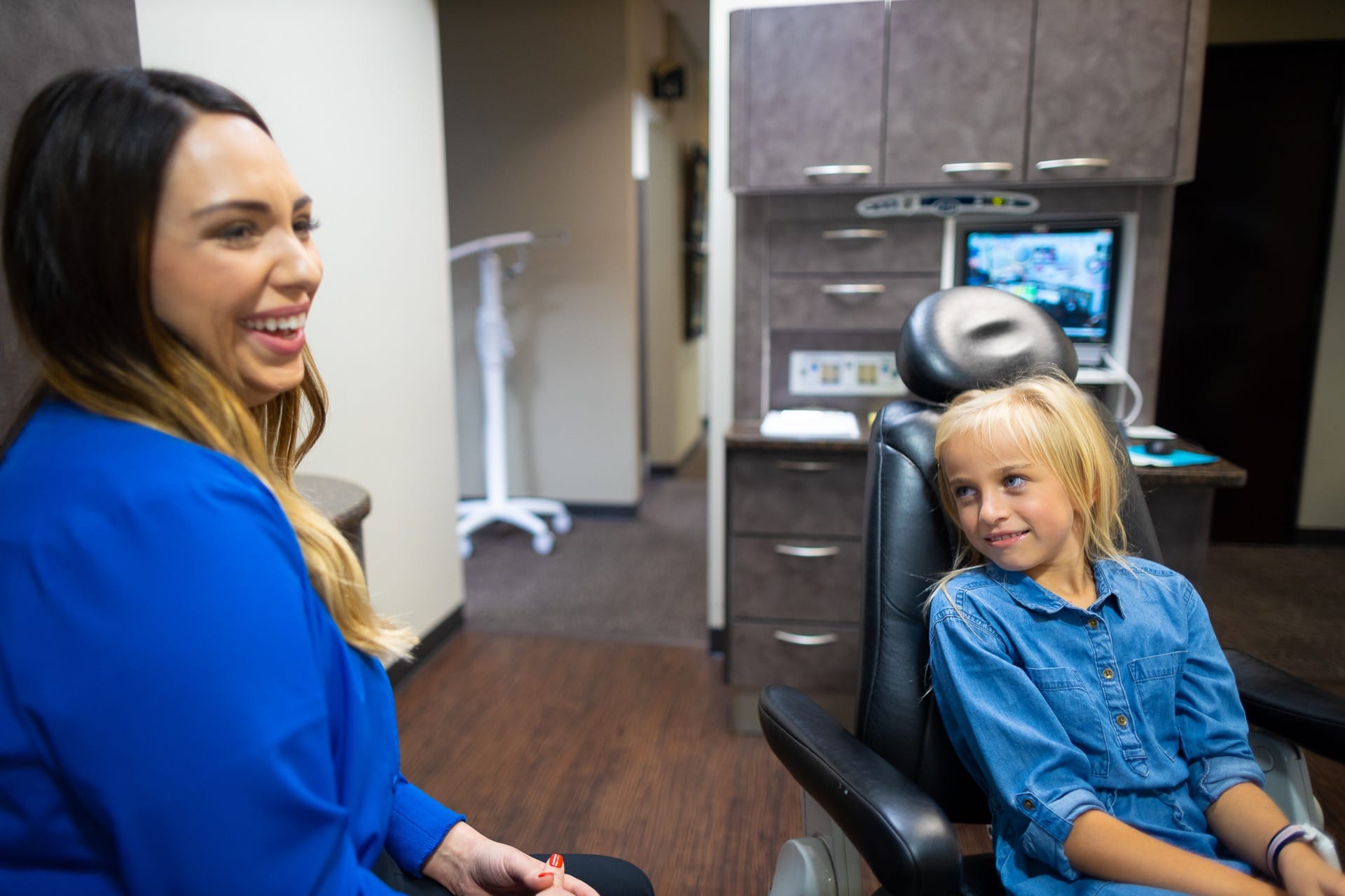 A dental professional in blue scrubs interacts warmly with a young patient in a modern dental office.