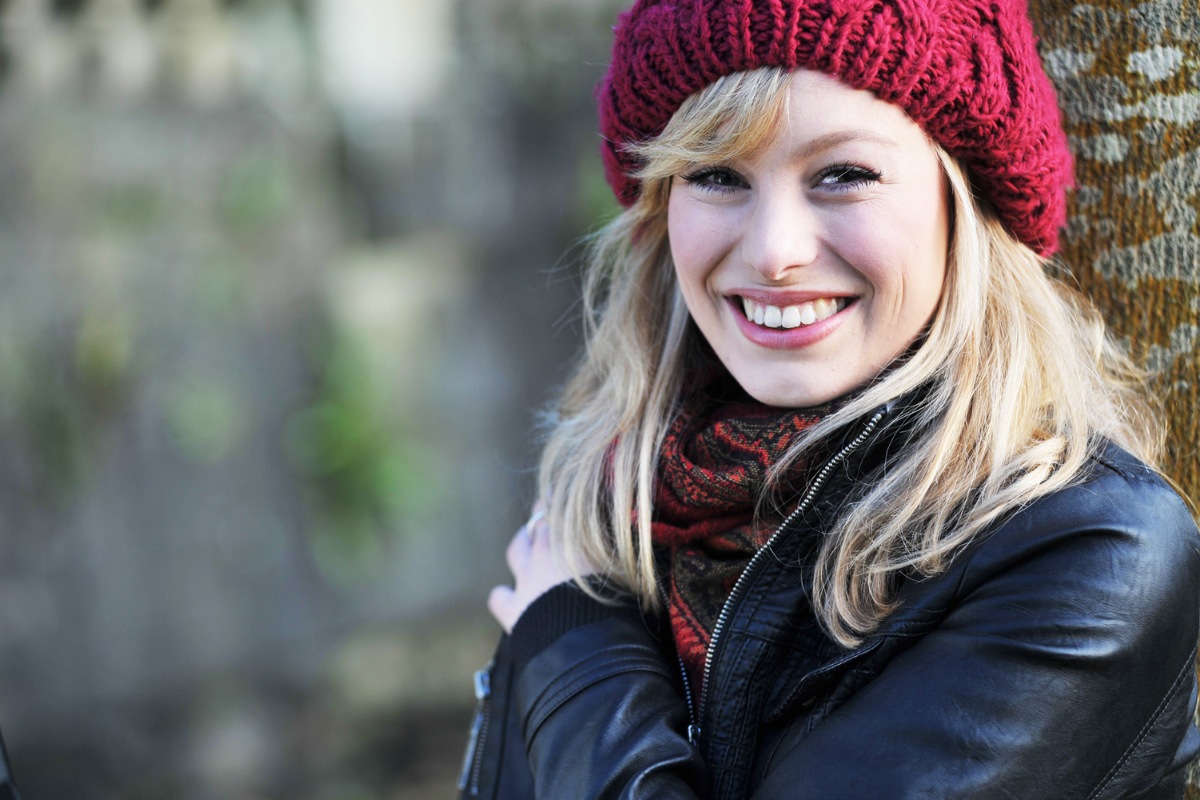 Smiling blonde woman wearing burgundy beanie and dark jacket outdoors in winter setting.