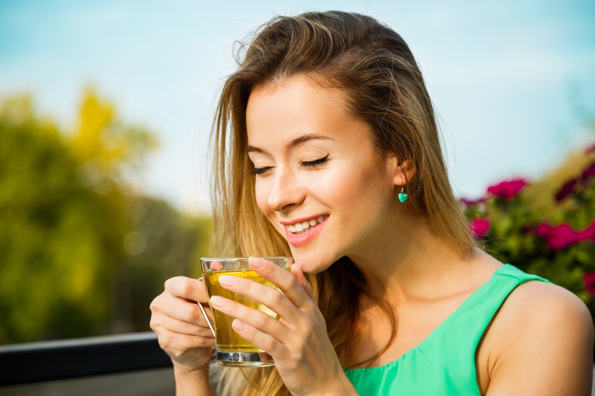 Woman in green top drinking water outdoors with garden and blue sky background.