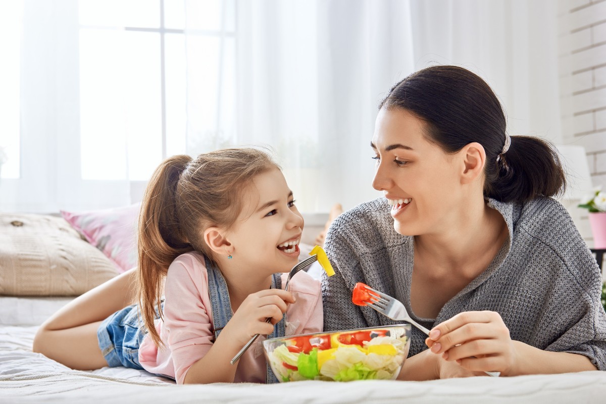 Mother and daughter eating fresh vegetables together on bed in bright bedroom.