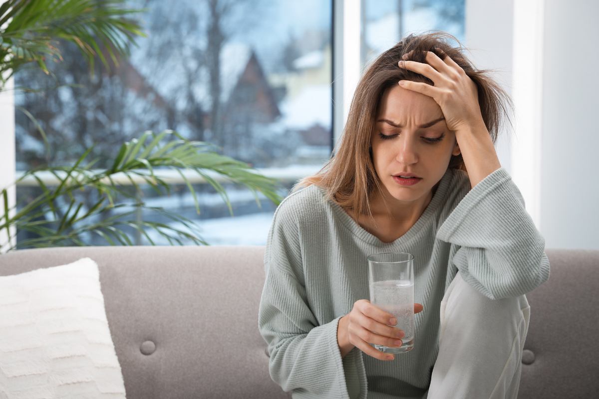 Woman at laptop holding water glass, hand to head, looking stressed in bright office by window.