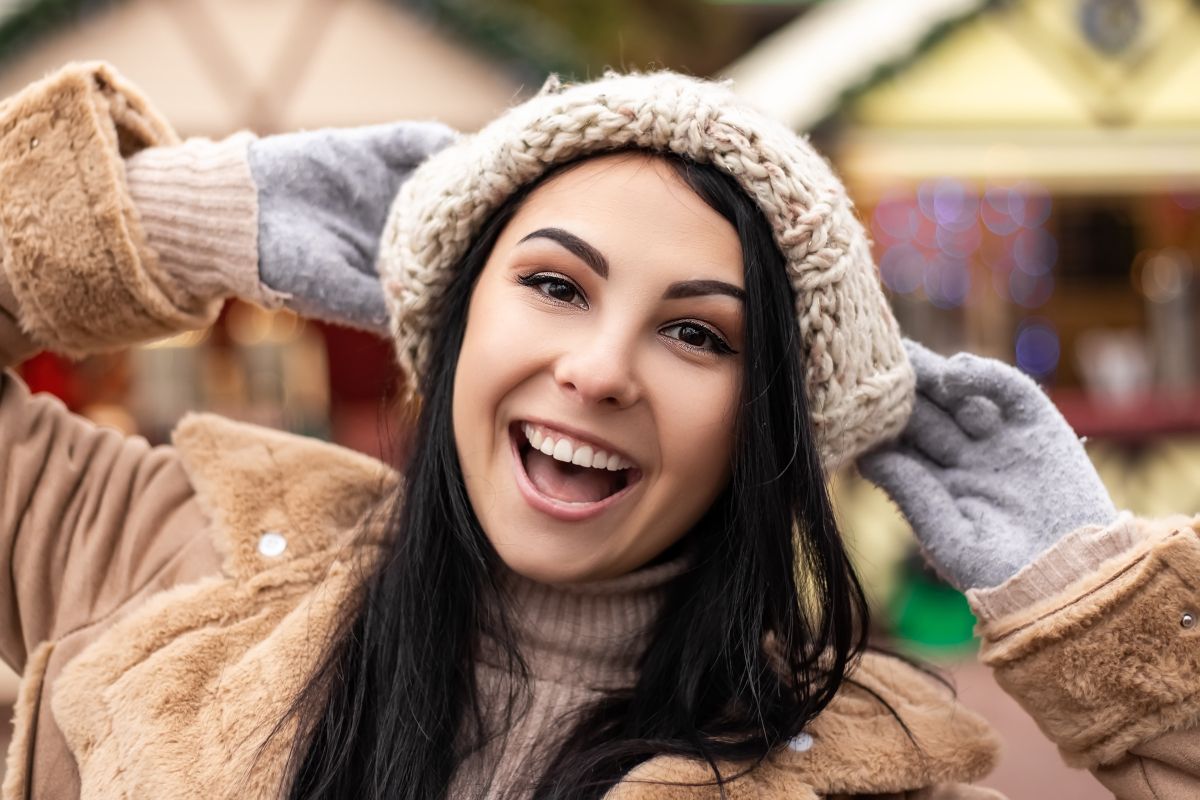 Woman wearing beige knit hat and tan jacket smiling outdoors at winter market.