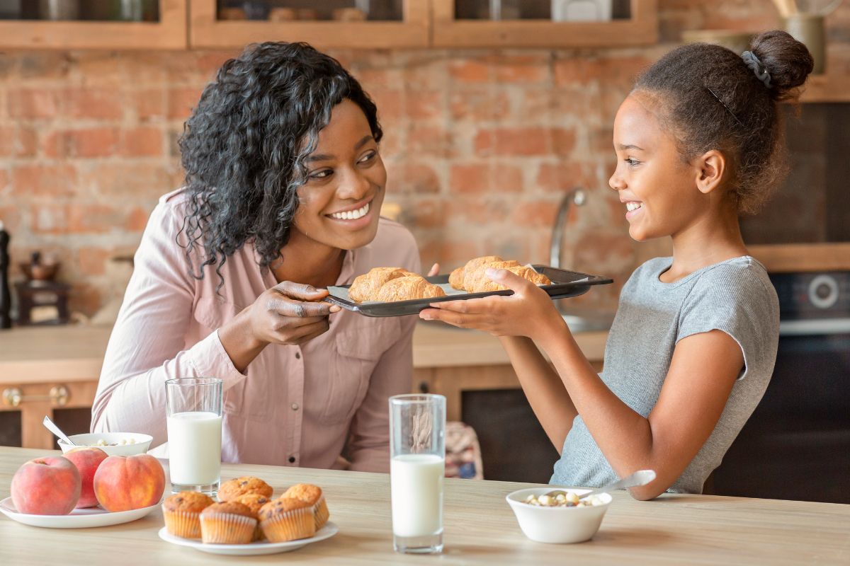 Woman and girl sharing breakfast pastries at kitchen table with brick backsplash.